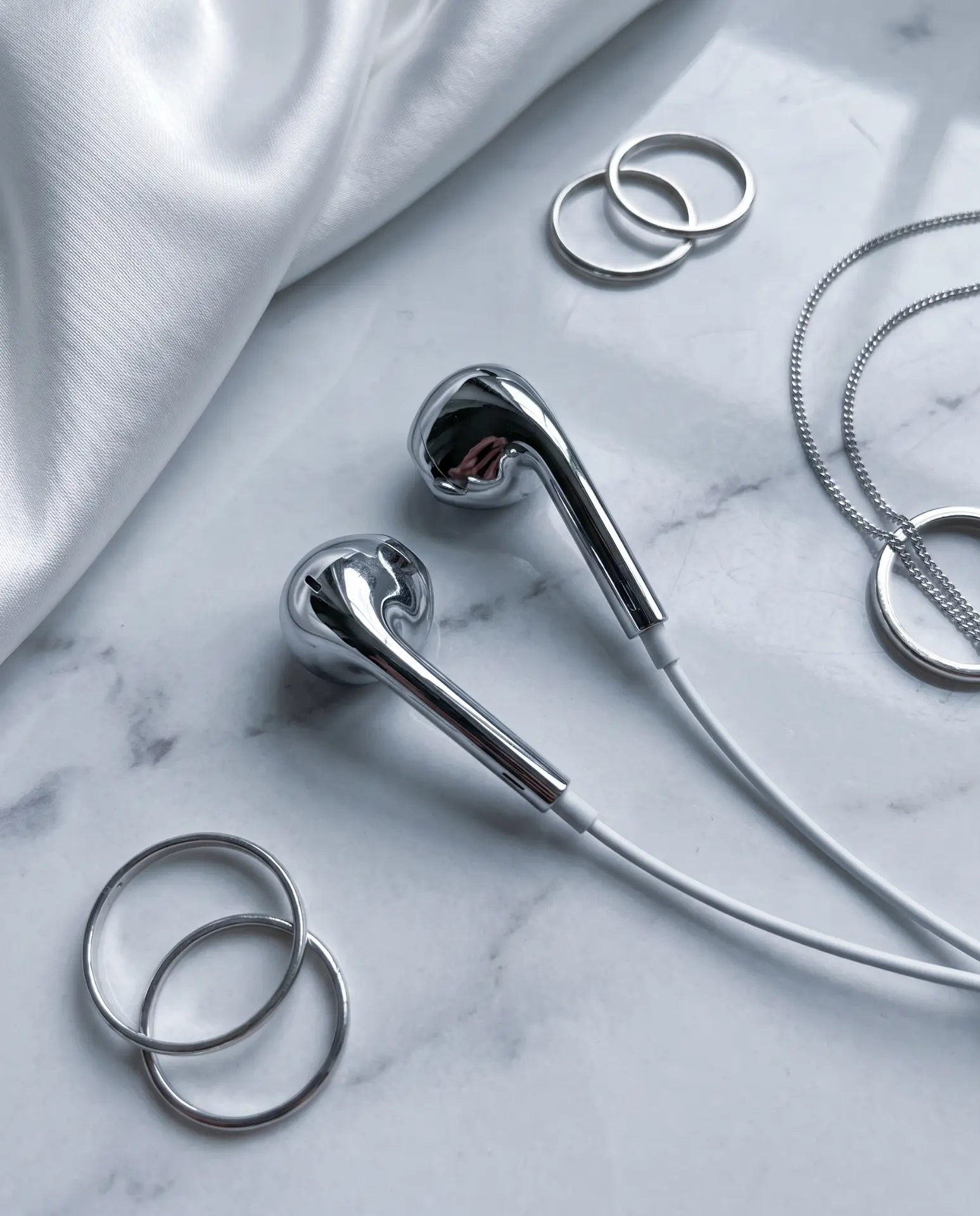 Silver chrome Auralie earbuds on marble with matching silver rings, chain necklace and white silk